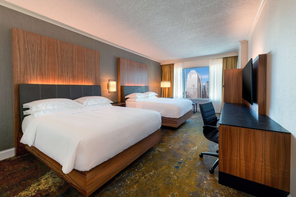 A hotel room with two beds, a desk, a chair, a TV, and a city view.