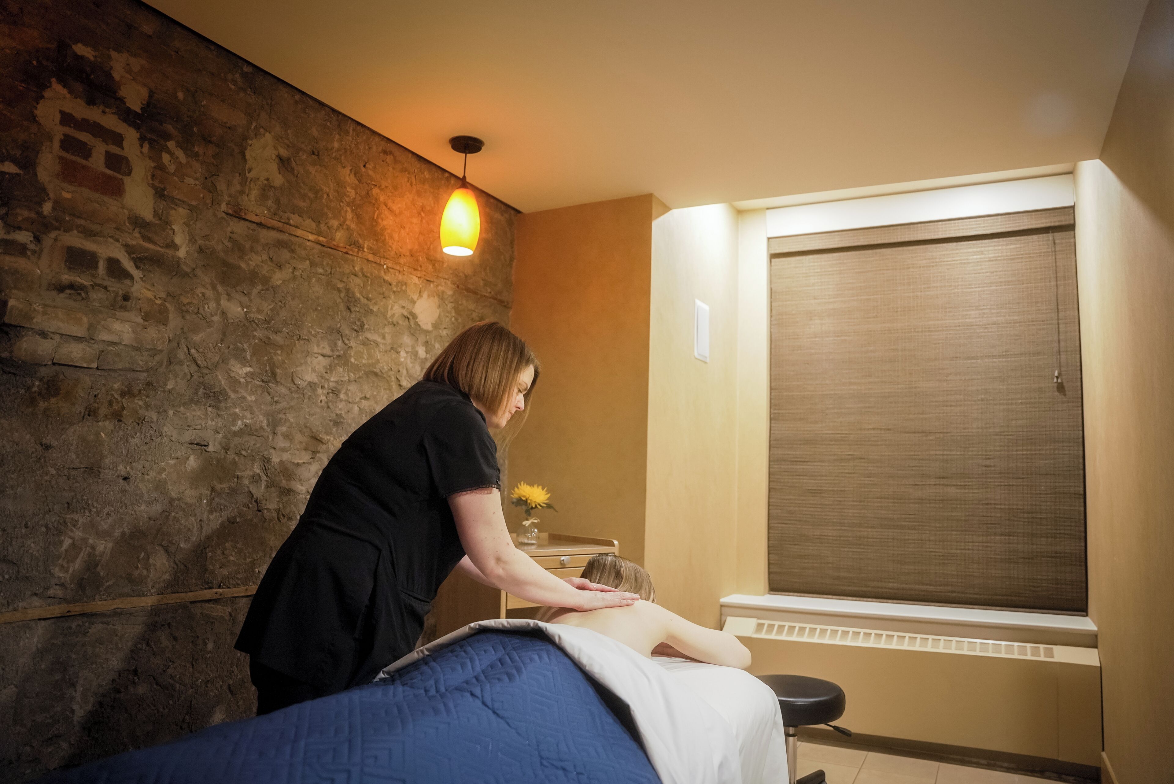 A massage therapy room with a brick wall, a wooden cabinet, and a window with blinds.