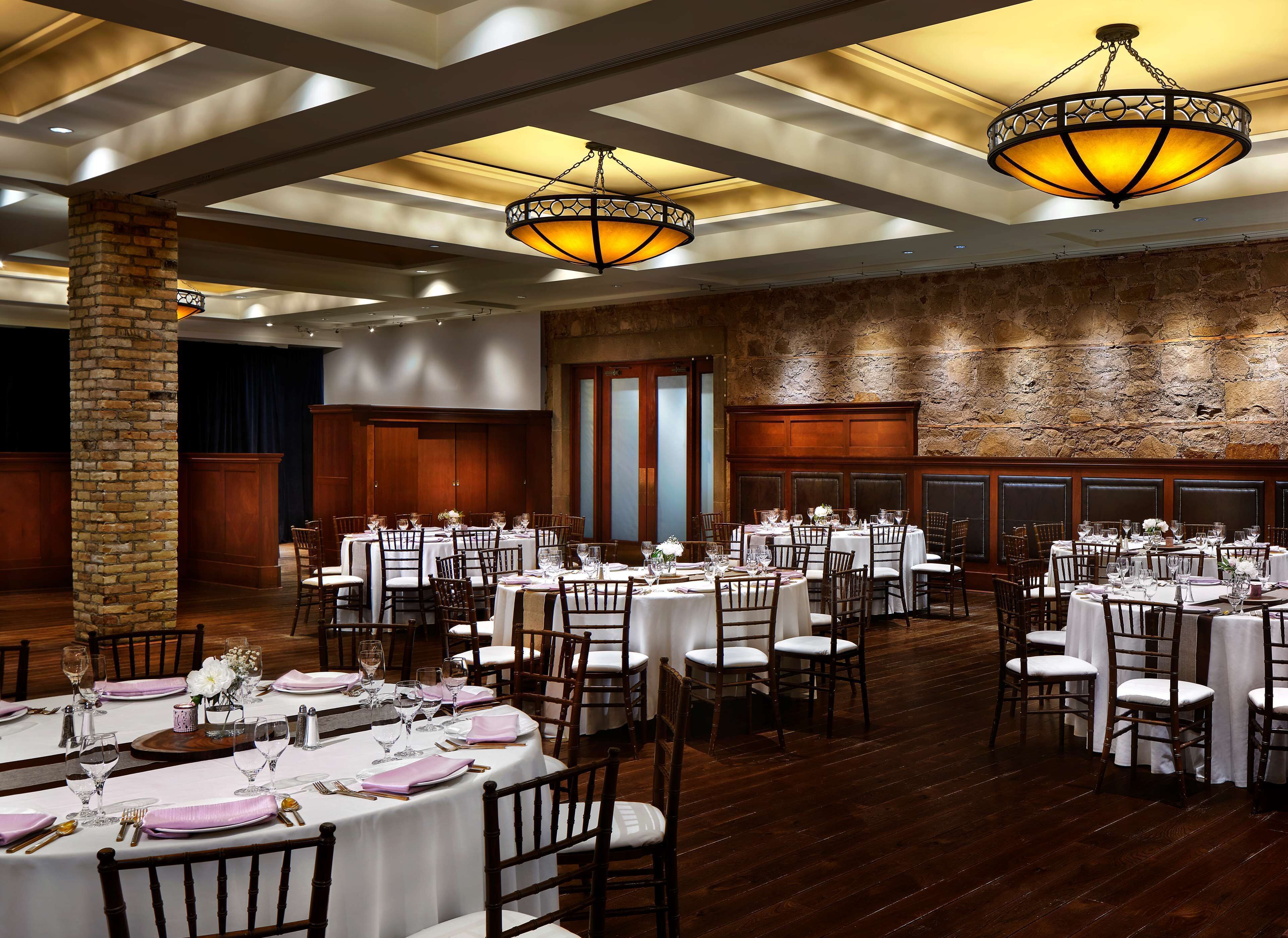 Une salle de banquet avec des tables rondes dressées pour le repas, des chaises en bois et un mur en pierre.