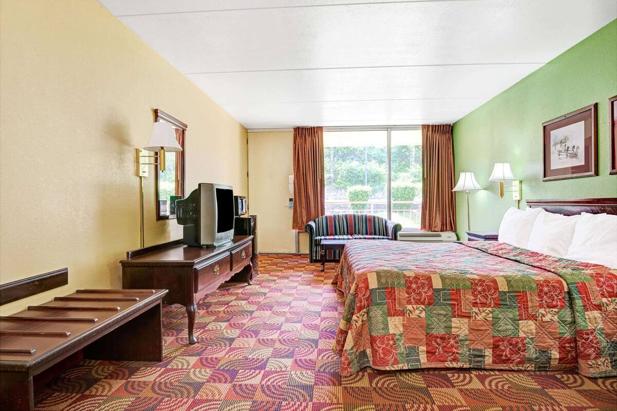 Une chambre d’hôtel avec un lit, un bureau avec une télévision, une chaise et une fenêtre avec des rideaux.