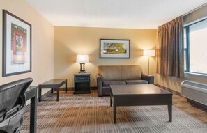 Habitación de hotel con sofá, una mesita, una silla, una lámpara, un cuadro en la pared y una ventana con cortinas.