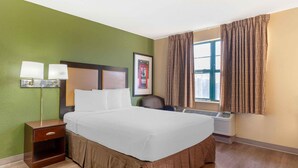 A hotel room with a bed, bedside lamps, a framed picture, a window with curtains, and an air conditioning unit.