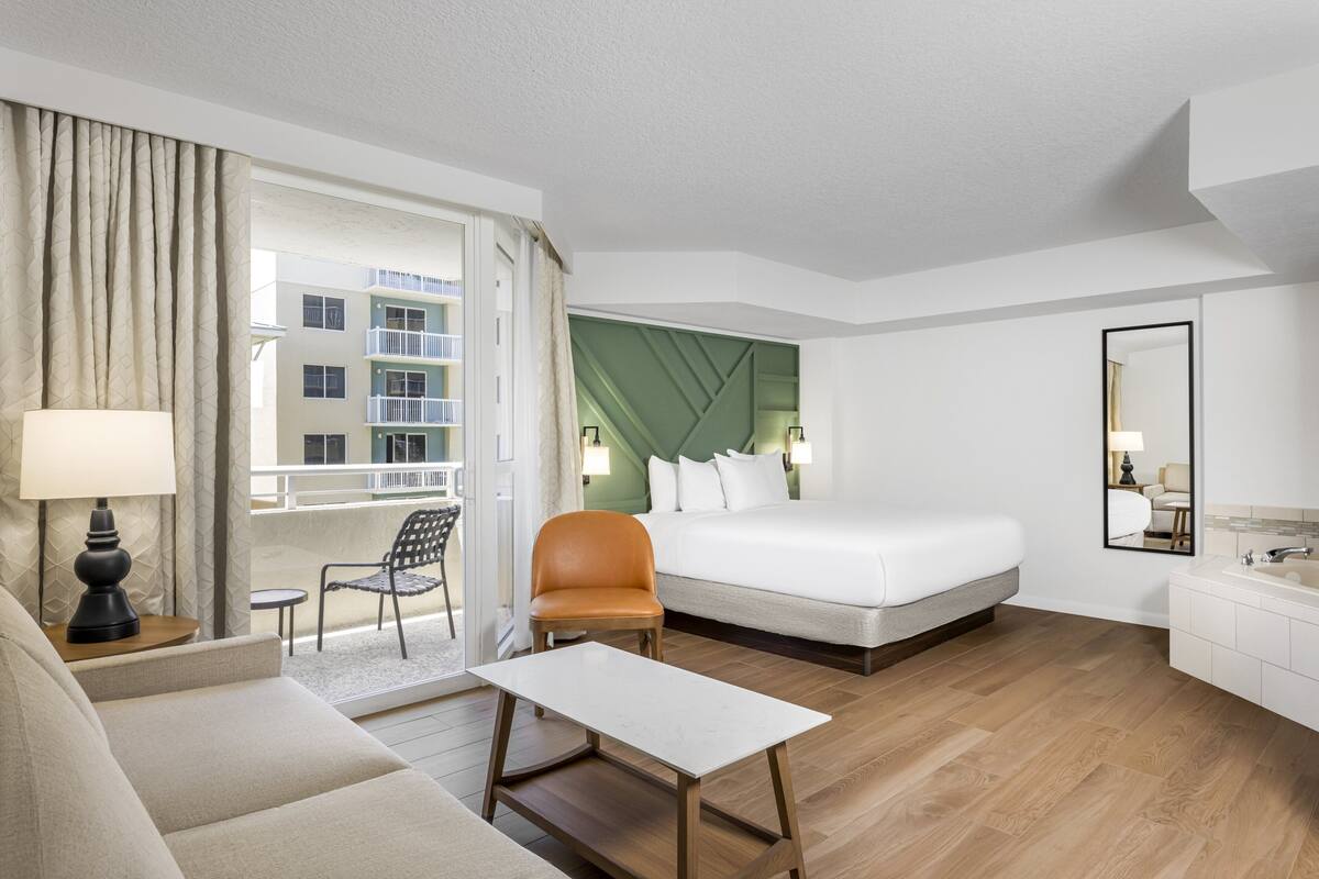 A hotel room with a bed, a sofa, a small table, a chair, a bathroom with a mirror, and a view of the outside building.