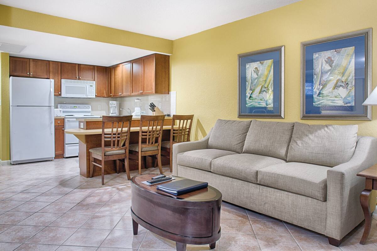 A living room with a beige sofa, a coffee table, and a dining area with wooden chairs and a table. There is a kitchen with wooden cabinets and a white refrigerator. Two framed paintings are on the wall.