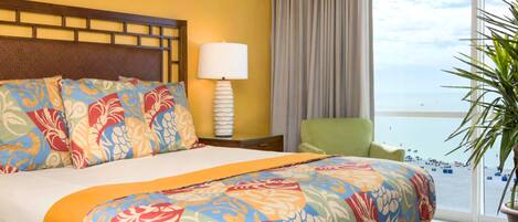 A hotel room with a bed, a nightstand with a lamp, a chair, a large window with a view of the beach, and a potted plant.