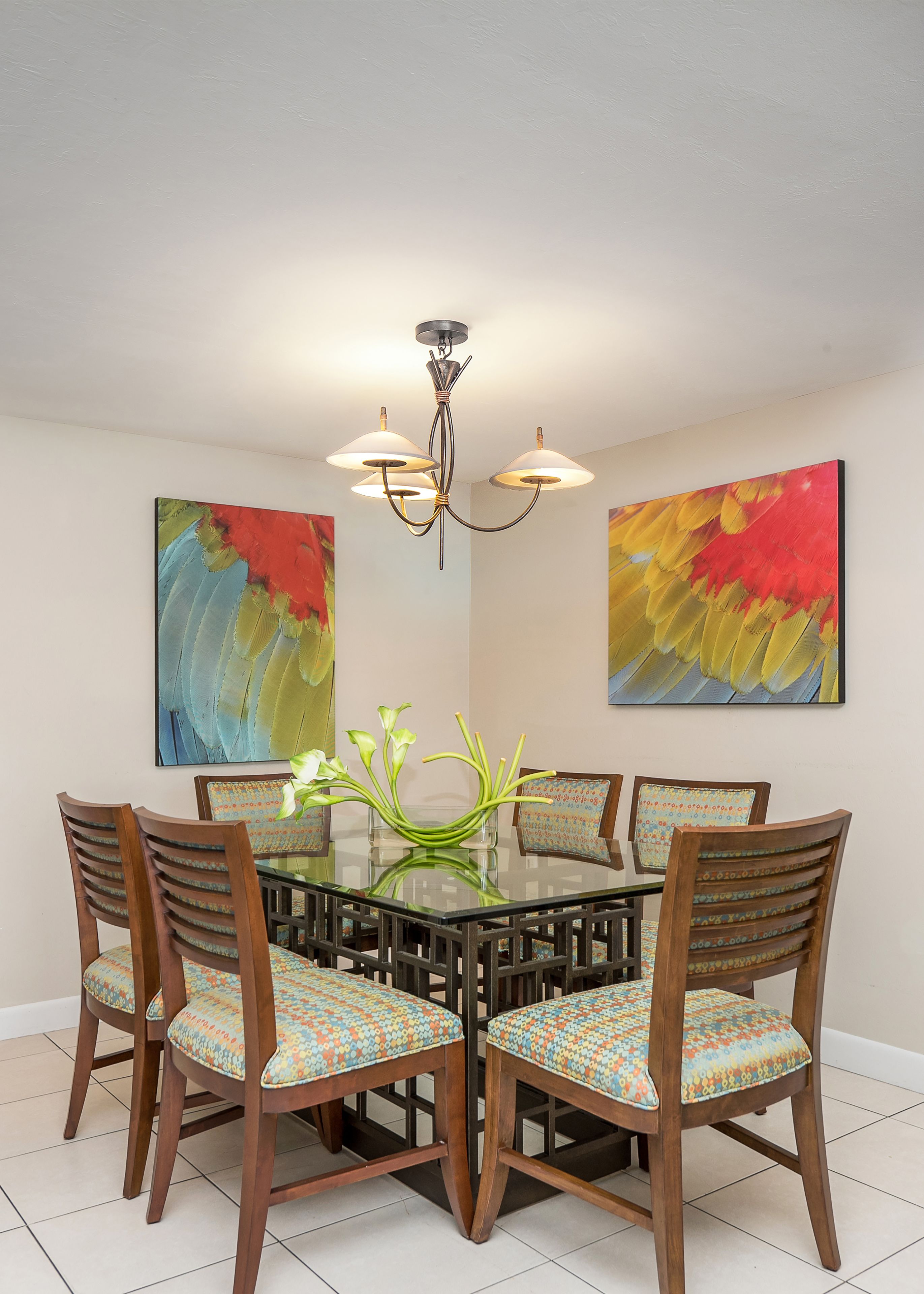 A dining area with a glass table surrounded by wooden chairs with patterned cushions, a chandelier, and abstract paintings on the walls.
