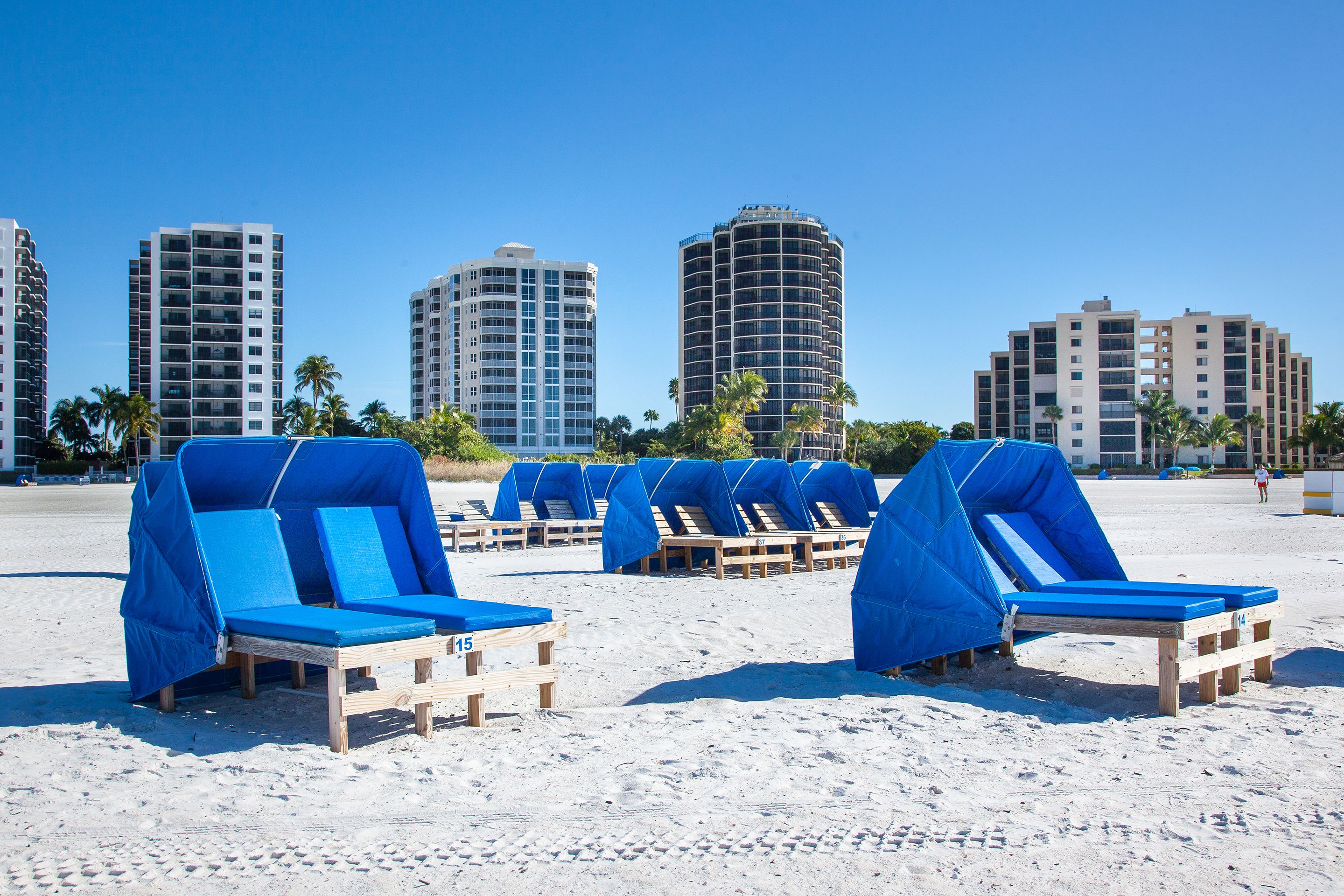 Beach chairs with blue covers, wooden loungers, and palm trees on a sandy beach with tall buildings in the background.