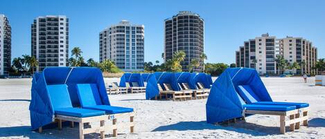 Beach chairs with blue covers, wooden loungers, and palm trees on a sandy beach with tall buildings in the background.
