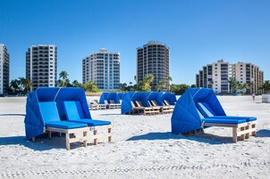 Beach chairs with blue covers, wooden loungers, and palm trees on a sandy beach with tall buildings in the background.