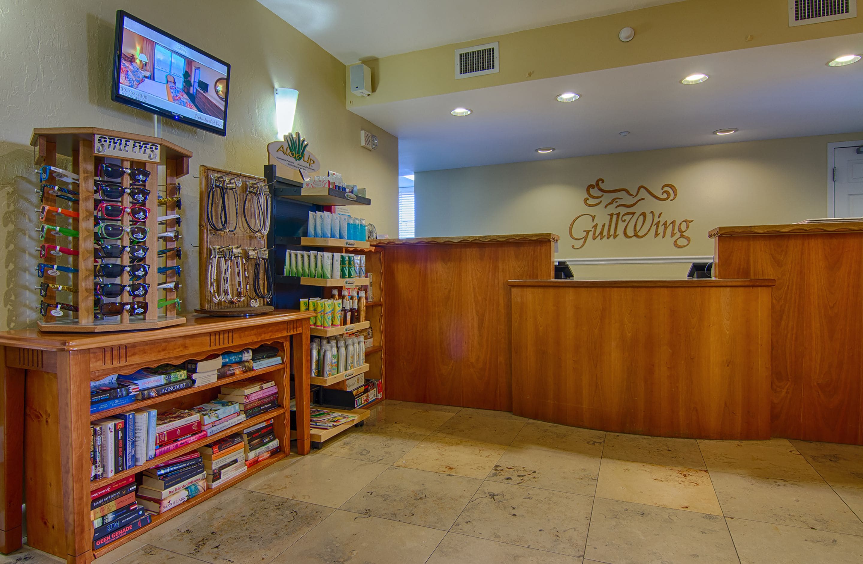 A reception area with a wooden desk, a TV mounted on the wall, and shelves with books and sunglasses.