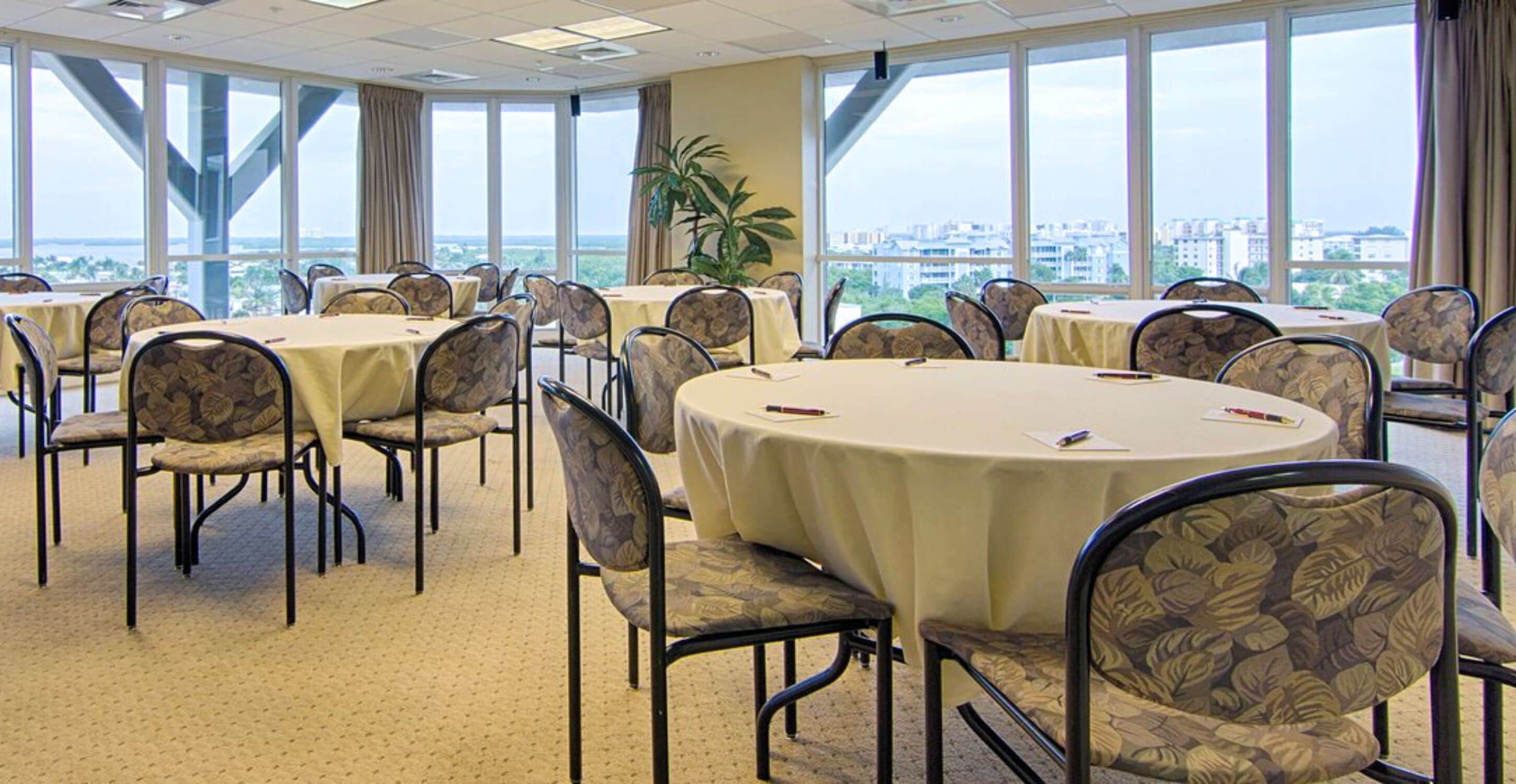 A conference room with round tables and chairs, large windows with a city view, and a potted plant.