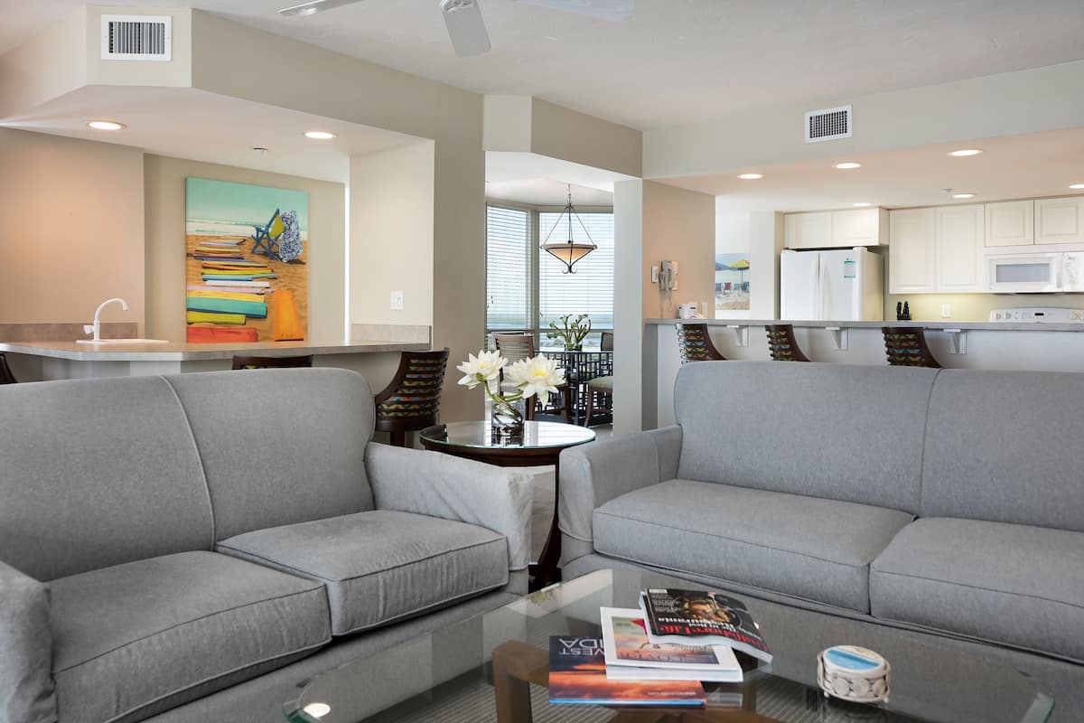 A modern living room with grey sofas, a glass coffee table, and a painting of a beach scene.
