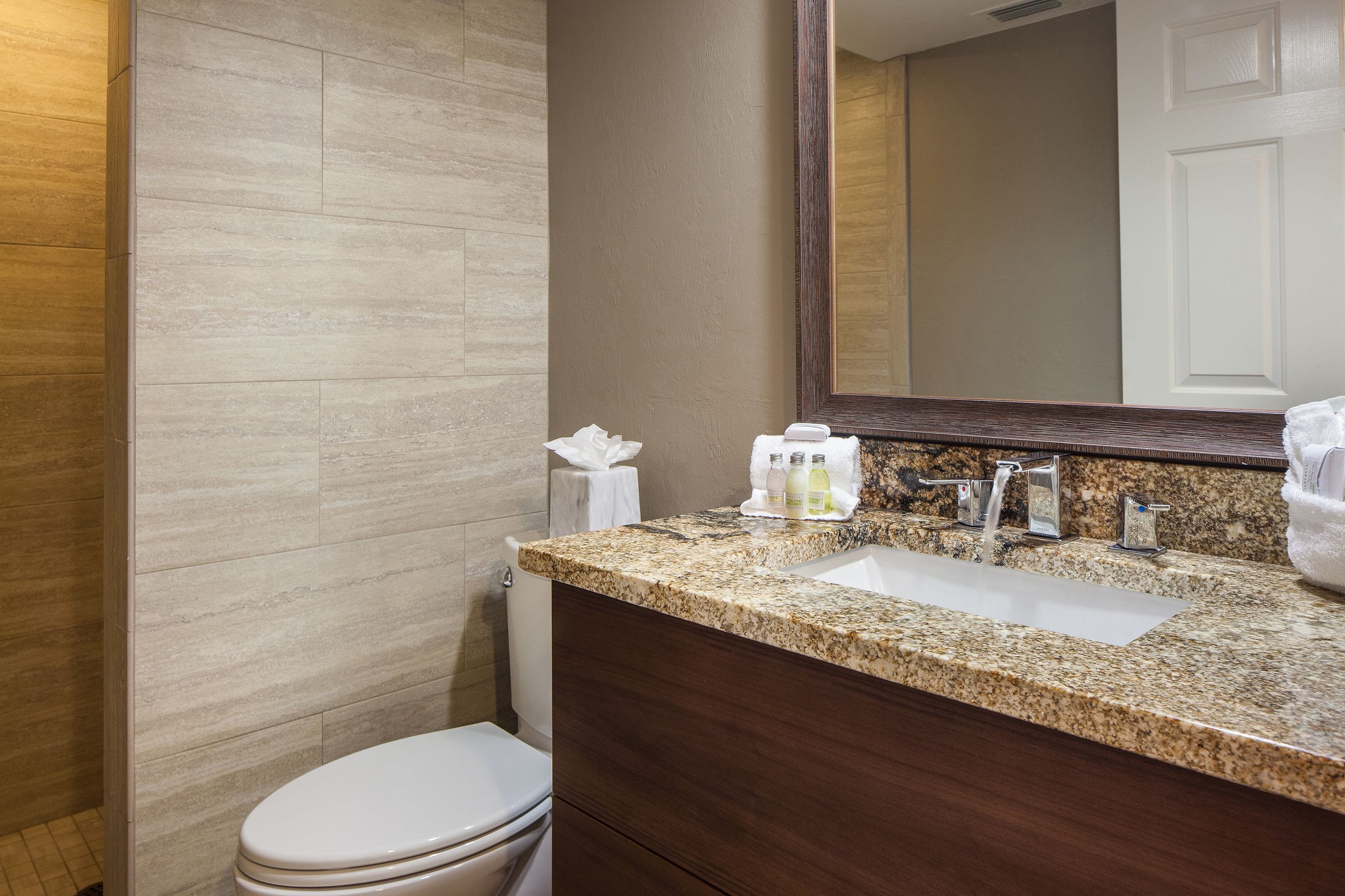A modern bathroom with a granite countertop, a white toilet, and a large mirror above the sink.