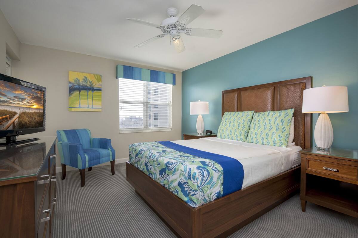 A hotel room with a bed, a desk, a chair, a TV, a ceiling fan, and a window with blinds.