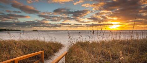 A wooden bench at the edge of a beach with a view of the sunset over the water.