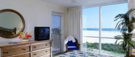 Ein Schlafzimmer mit Bett, Deckenventilator, Kommode, Fernseher und einem großen Fenster mit Meerblick.