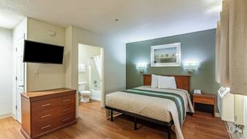 A hotel room with a bed, bedside tables, a dresser, a flat-screen TV, a painting on the wall, and a bathroom visible through an open door.