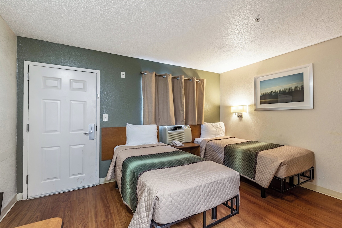 Habitación de hotel con dos camas, una puerta, equipo de aire acondicionado, una mesita de noche, una lámpara y un cuadro en la pared.