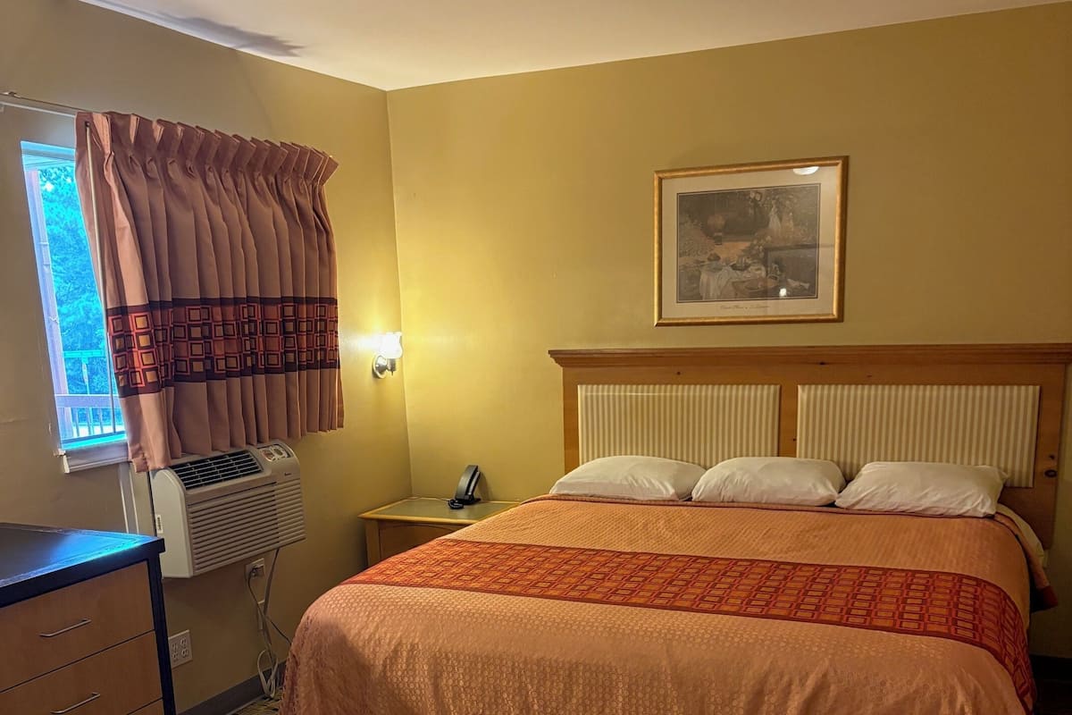 A hotel room with a bed, a desk, a chair, a radiator, and a window with curtains.