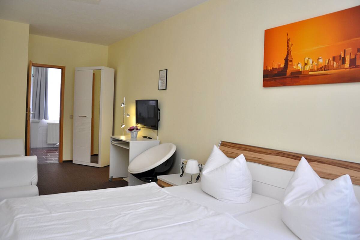 A hotel room with a bed, a desk with a chair, a television, and a painting of a cityscape.