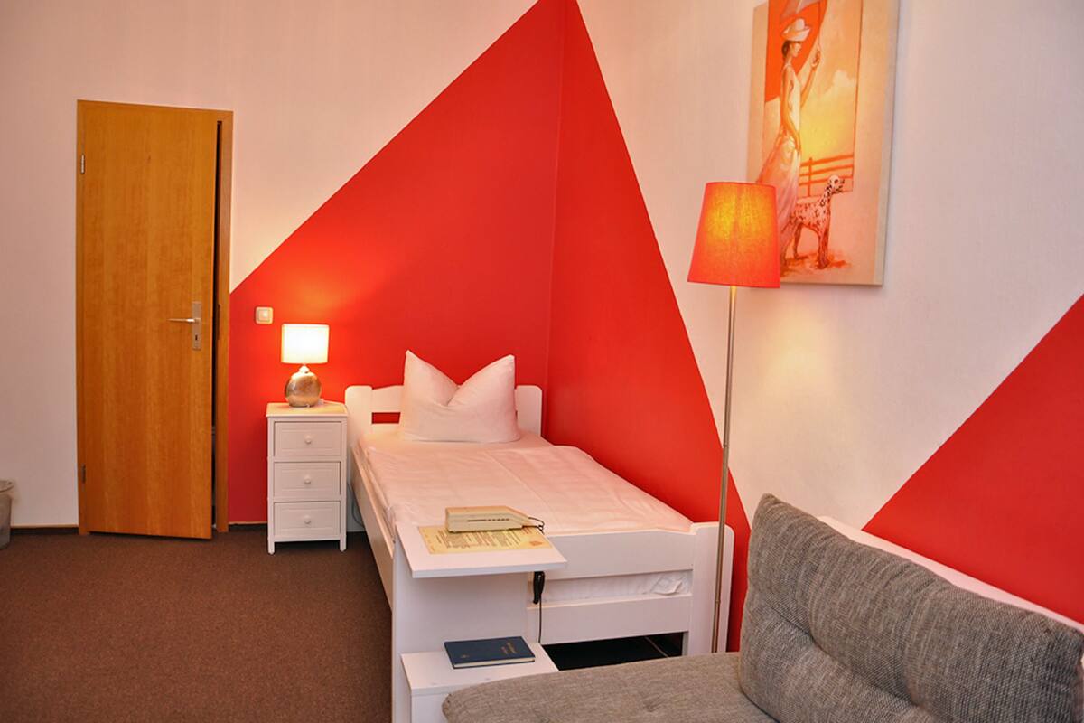 A small, single-bed room with a red and white color scheme, a bedside table with a lamp, and a painting on the wall.