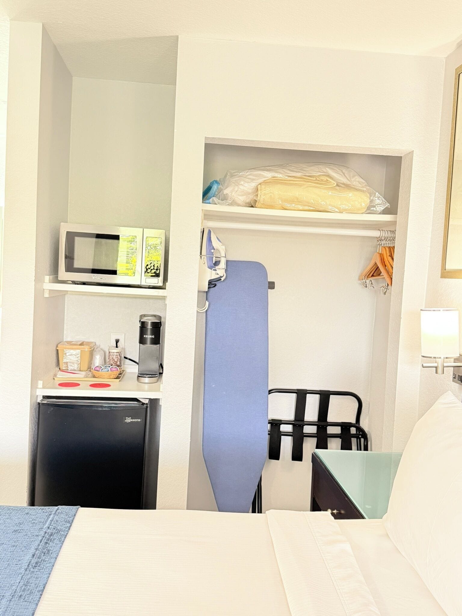 Habitació d'hotel amb un llit, un petit escriptori amb microones, cafetera i nevera miniatura. Hi ha un armari amb roba penjada i una tovallola blava.
