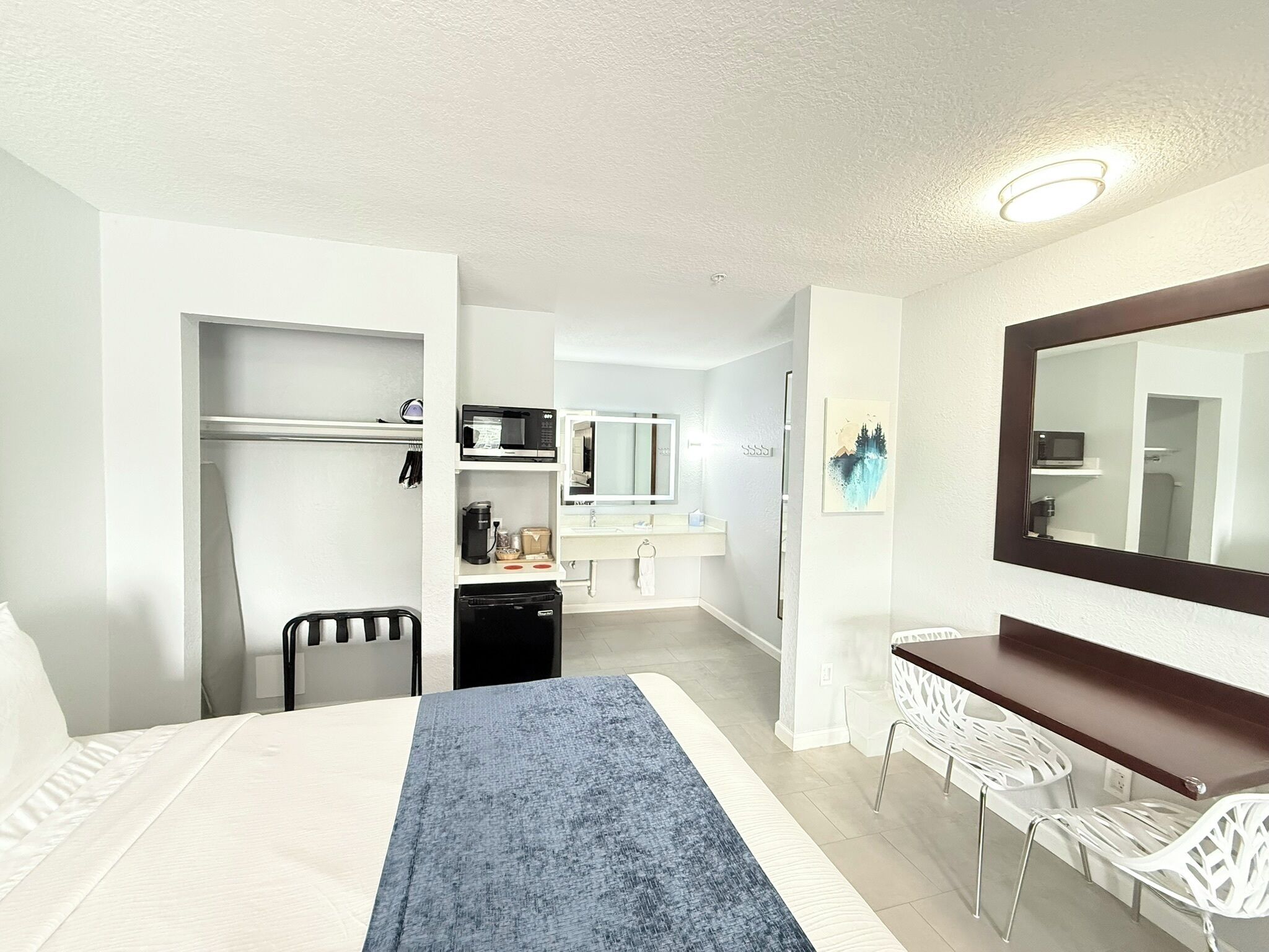 A hotel room with a bed, a desk, a chair, a microwave, a coffee maker, a mirror, and a bathroom in the background.