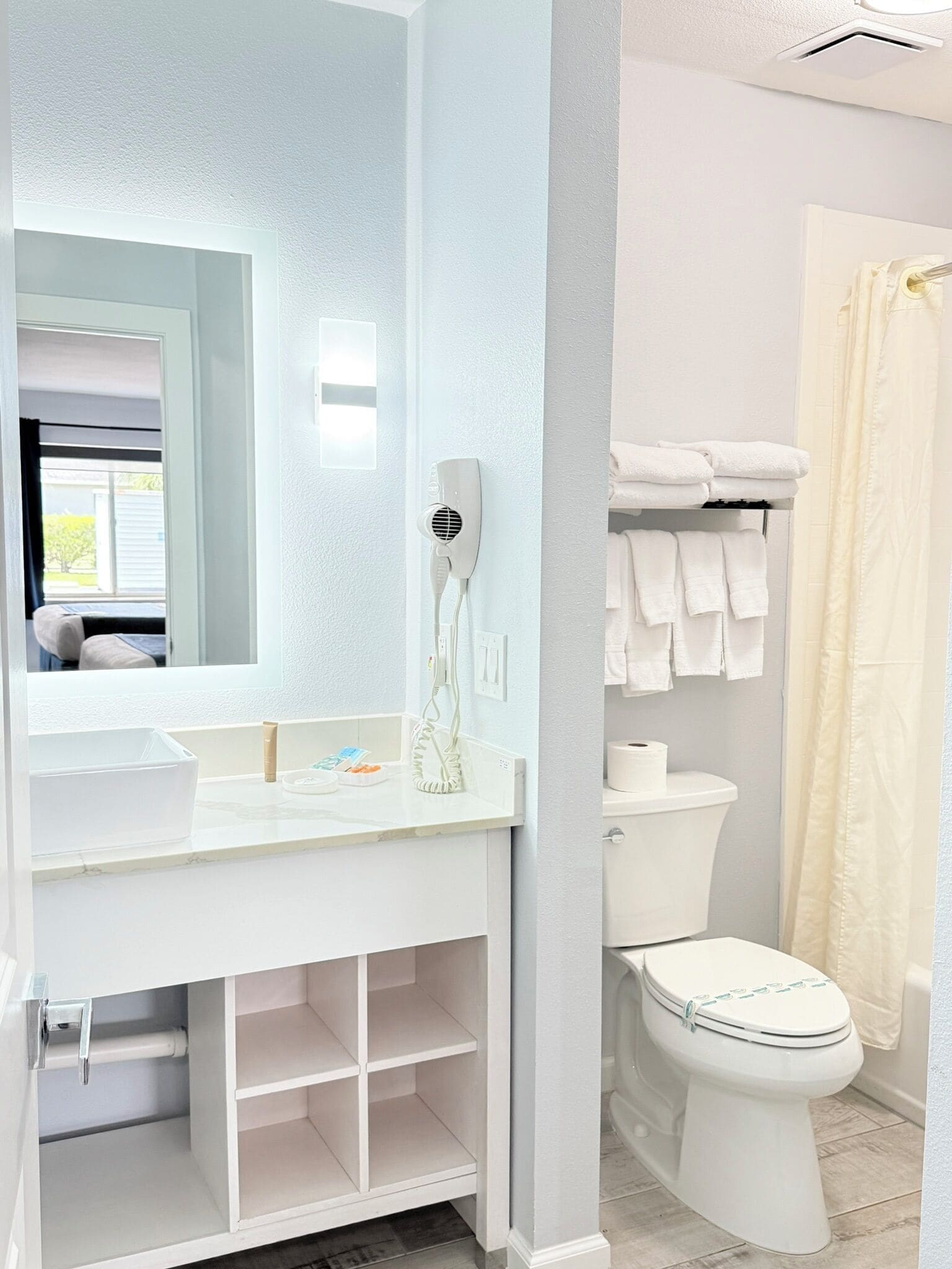 A bathroom with a white sink, mirror, and toilet. There is a towel rack with folded towels and a shower curtain.