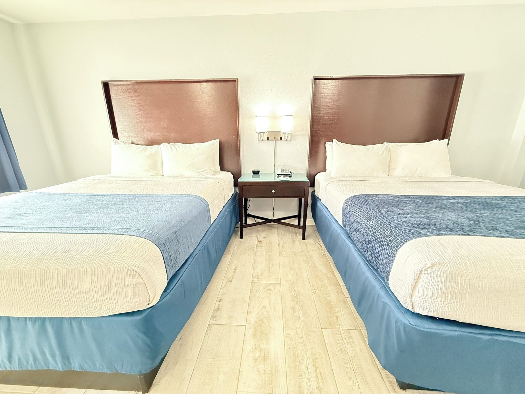 A hotel room with two beds, a nightstand, a lamp, and a wooden floor.
