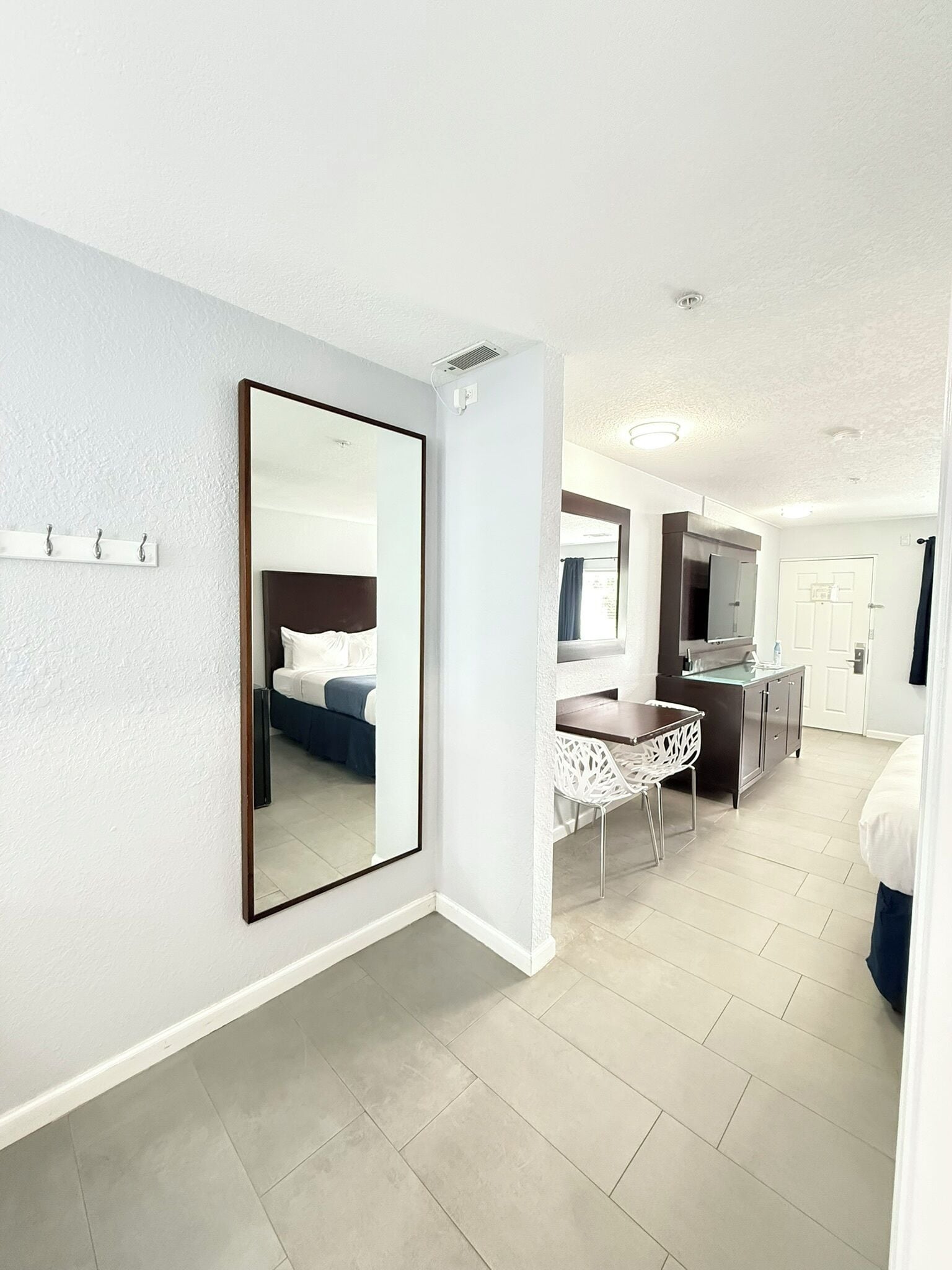A hotel room with a large mirror, a bed, a desk with a chair, and a kitchenette.
