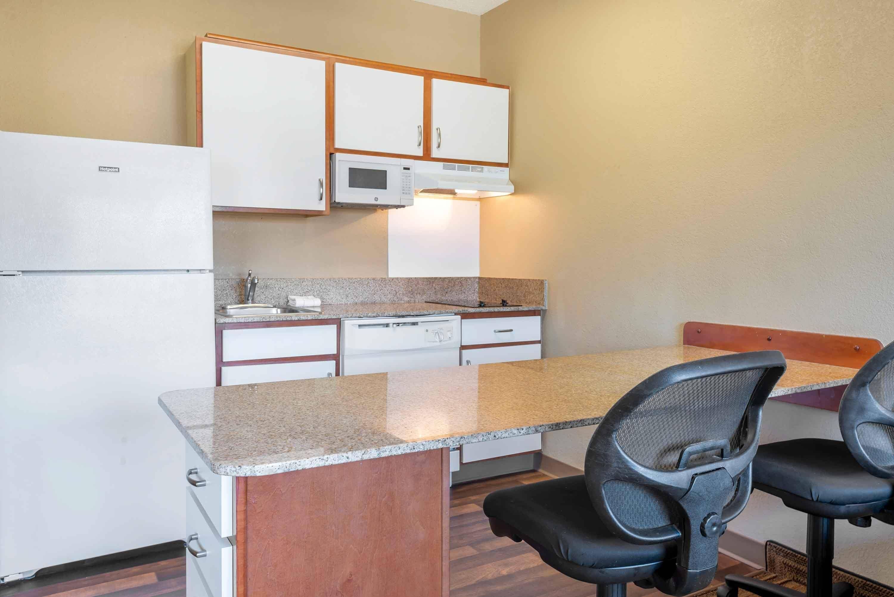 A compact kitchen with a refrigerator, microwave, and dishwasher, a granite countertop, and two black office chairs.
