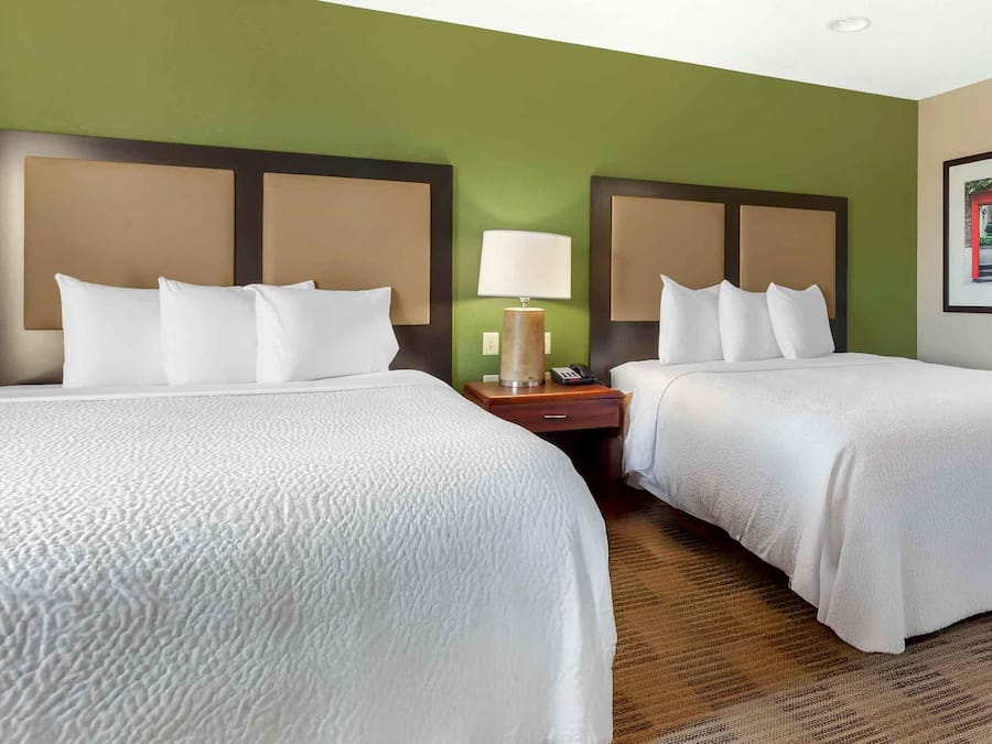 Quarto de hotel com duas camas, uma mesinha de cabeceira, um abajur e um quadro na parede.