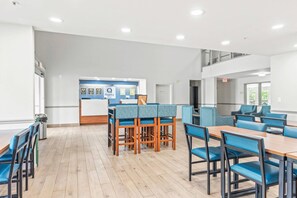 Un comedor amplio con mesas de madera y sillas tapizadas en azul, una recepción con diplomas enmarcados y un techo alto con iluminación empotrada.