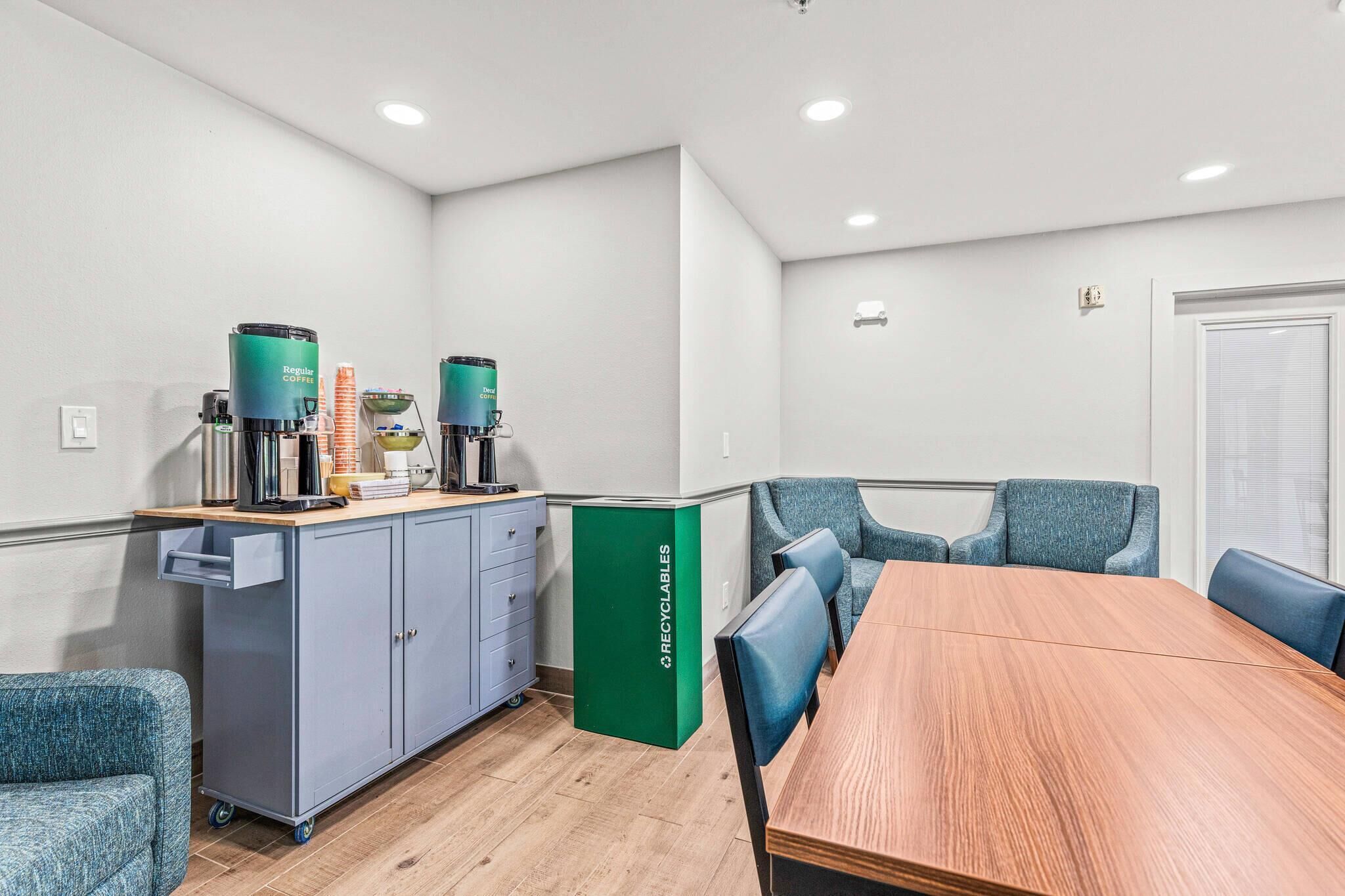 A conference room with a wooden table, blue chairs, a grey cabinet with a recycling bin, and two coffee machines.