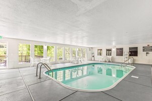 Piscina interior com paredes de vidro transparente, cadeiras brancas e posto de salva-vidas.