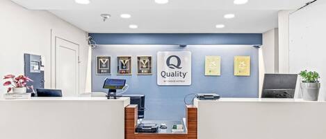 Hotellin vastaanottotiski, jonka takana on sininen seinä, seinälle ripustettuja palkintoja, kyltti, jossa lukee ”Quality by Choice Hotels”, tietokone ja kahvinkeitin.