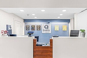 Uma receção de hotel com parede azul, prêmios emoldurados, um sinal de 'Quality by Choice Hotels', um computador e uma máquina de café.