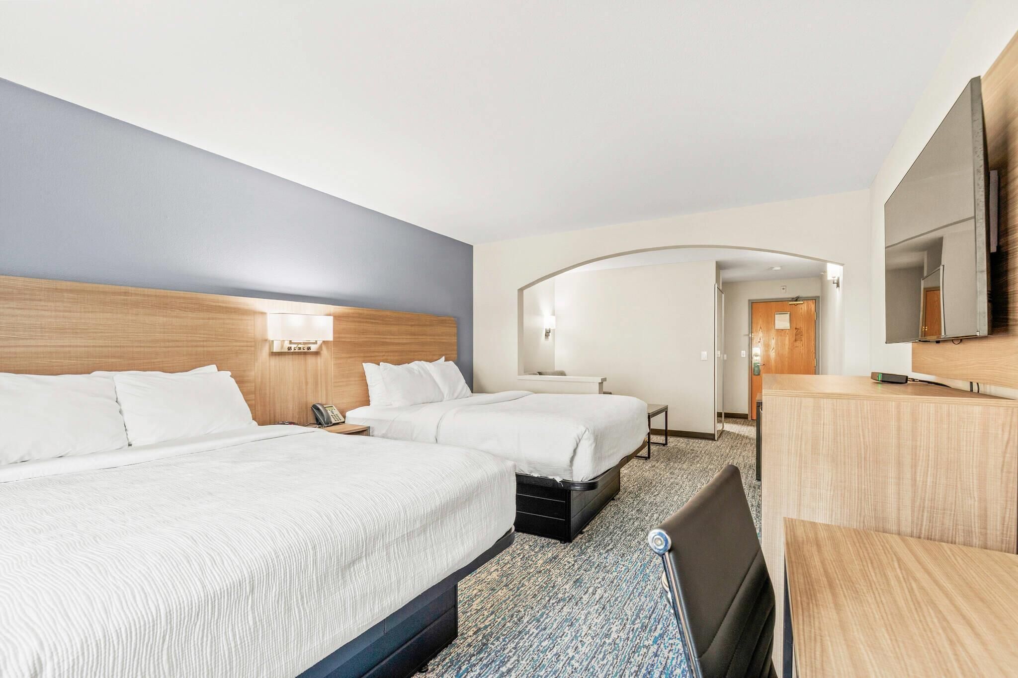 Habitación de hotel con dos camas, un escritorio, una silla, un televisor y un baño visible a través de un arco.