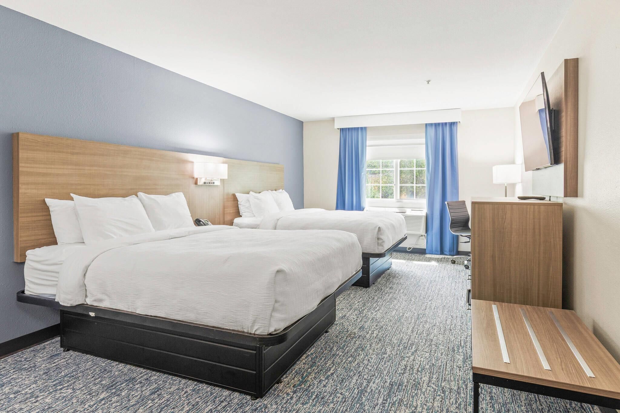 A hotel room with two beds, a desk, a chair, a TV, and a window with curtains.
