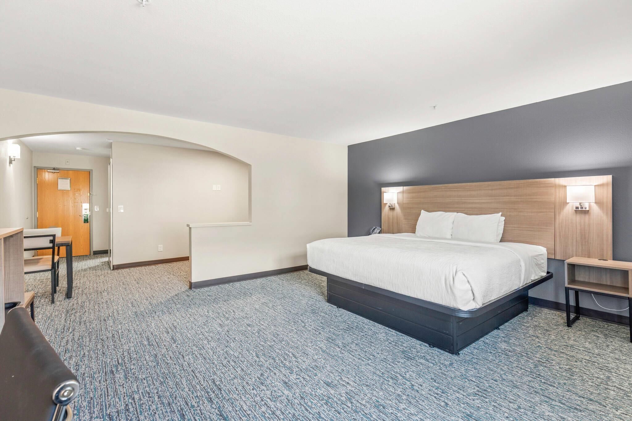 A hotel room with a large bed, a desk, and a chair. There is a door leading to another room and a small table with a lamp.