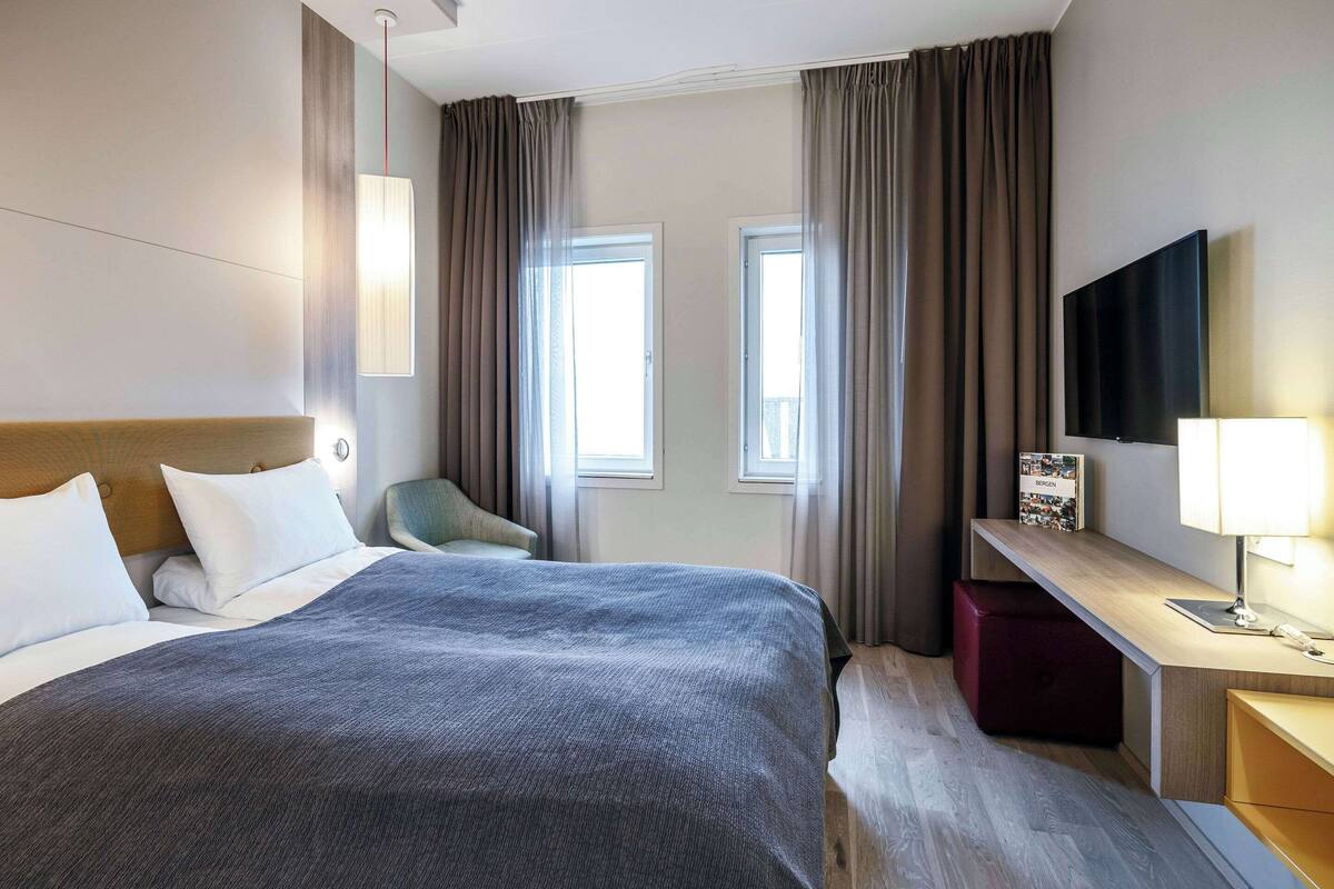Ein Hotelzimmer mit Bett, Schreibtisch, Sessel, Fernseher und zwei Fenstern mit Vorhängen.