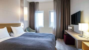 Una habitación de hotel con cama, escritorio, silla, televisor y dos ventanas con cortinas.