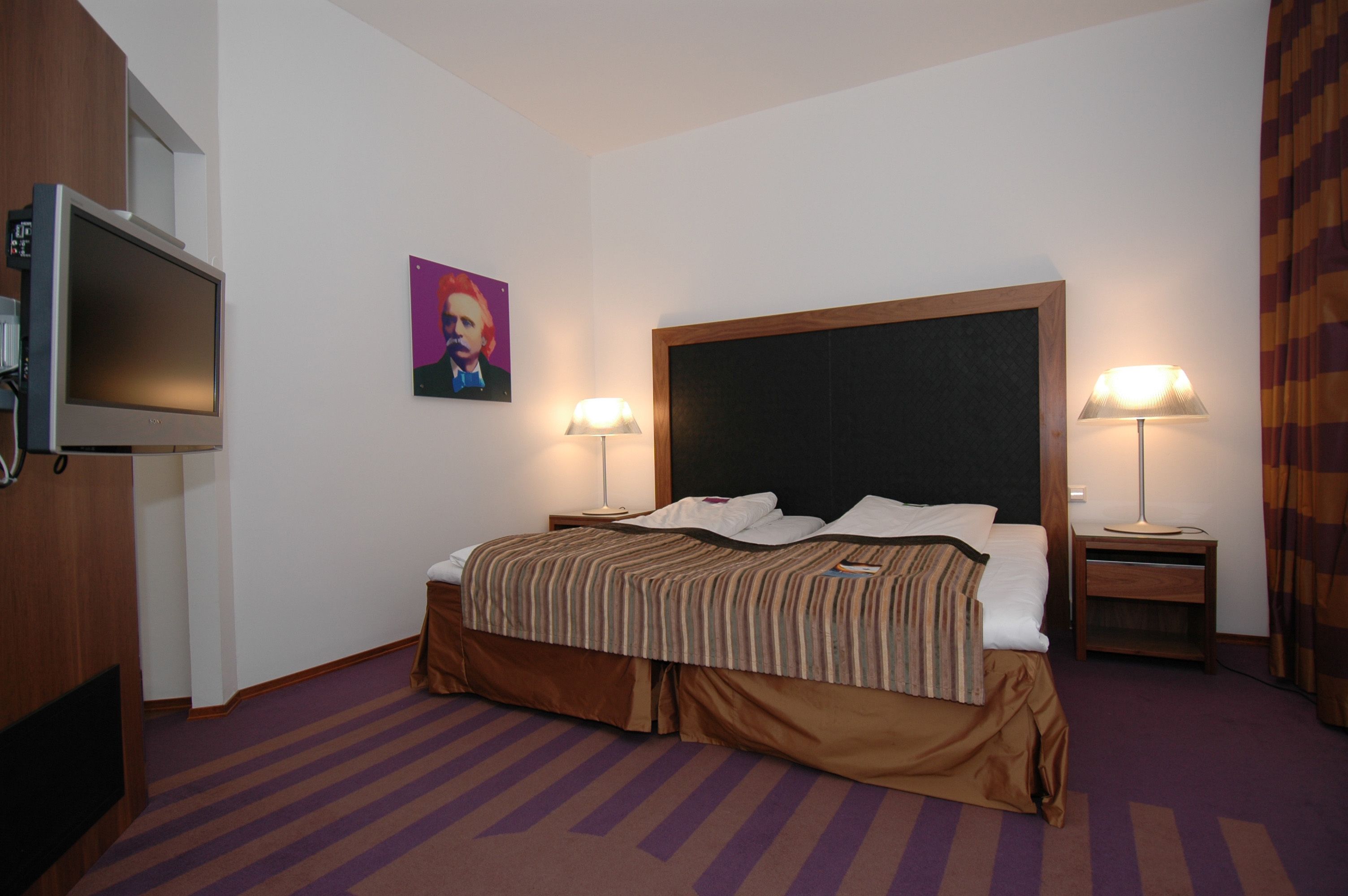 Una camera d'albergo con un letto, due lampade ai comodini, una televisione, un quadro e un poggiatesta in legno.