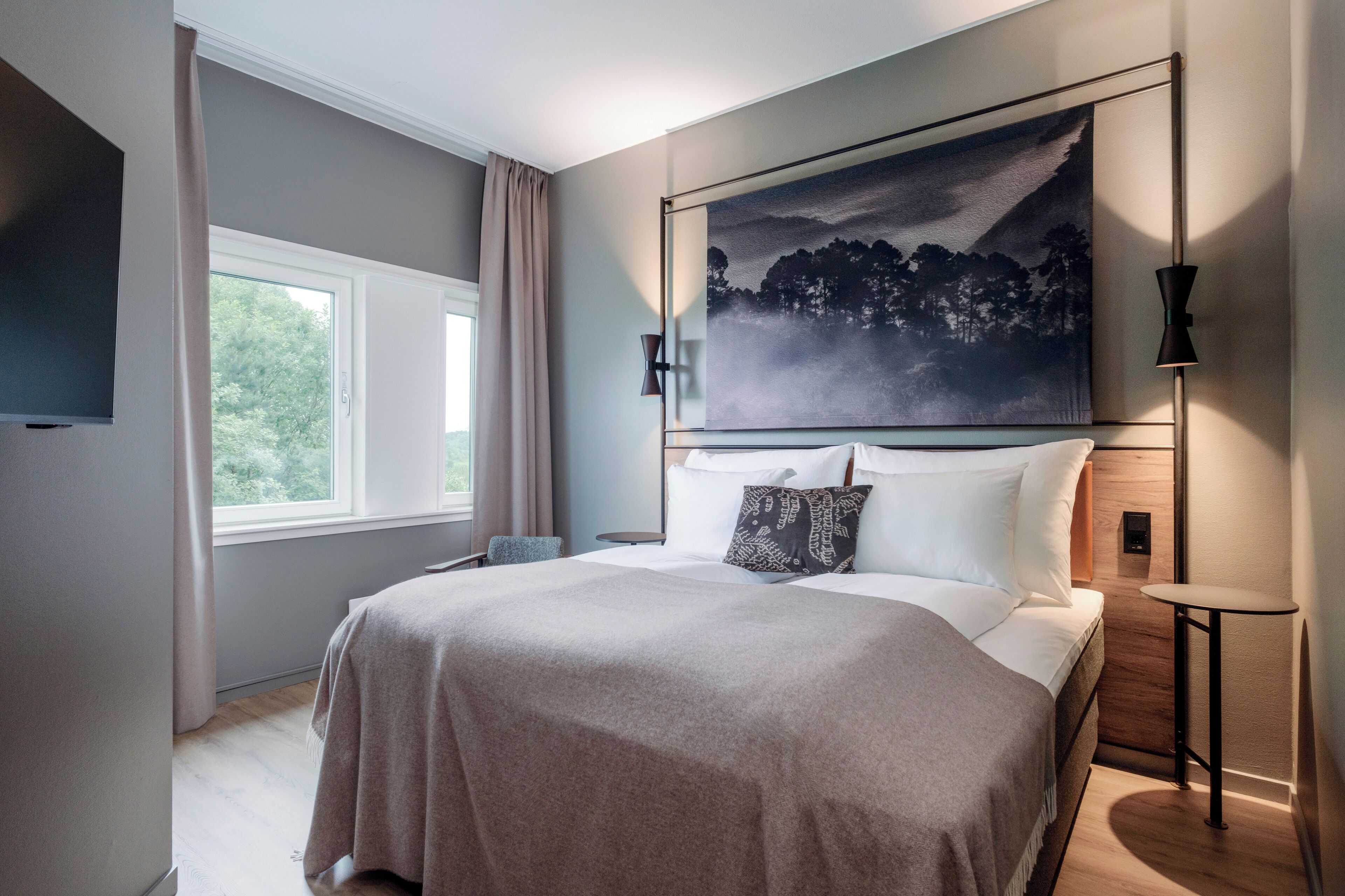 A modern hotel room with a large bed, a flat-screen TV, a painting above the bed, and a small table by the window.