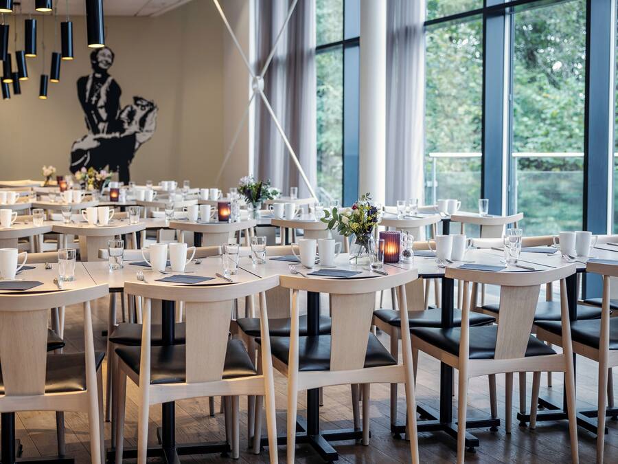A modern restaurant with round tables and black leather chairs, large windows, and a mural on the wall.