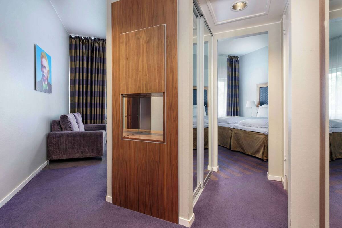 Ein Hotelzimmer mit einer Holzwand, einem Sofa, einem Bett mit Kopfteil und einem großen Fenster mit Vorhängen.