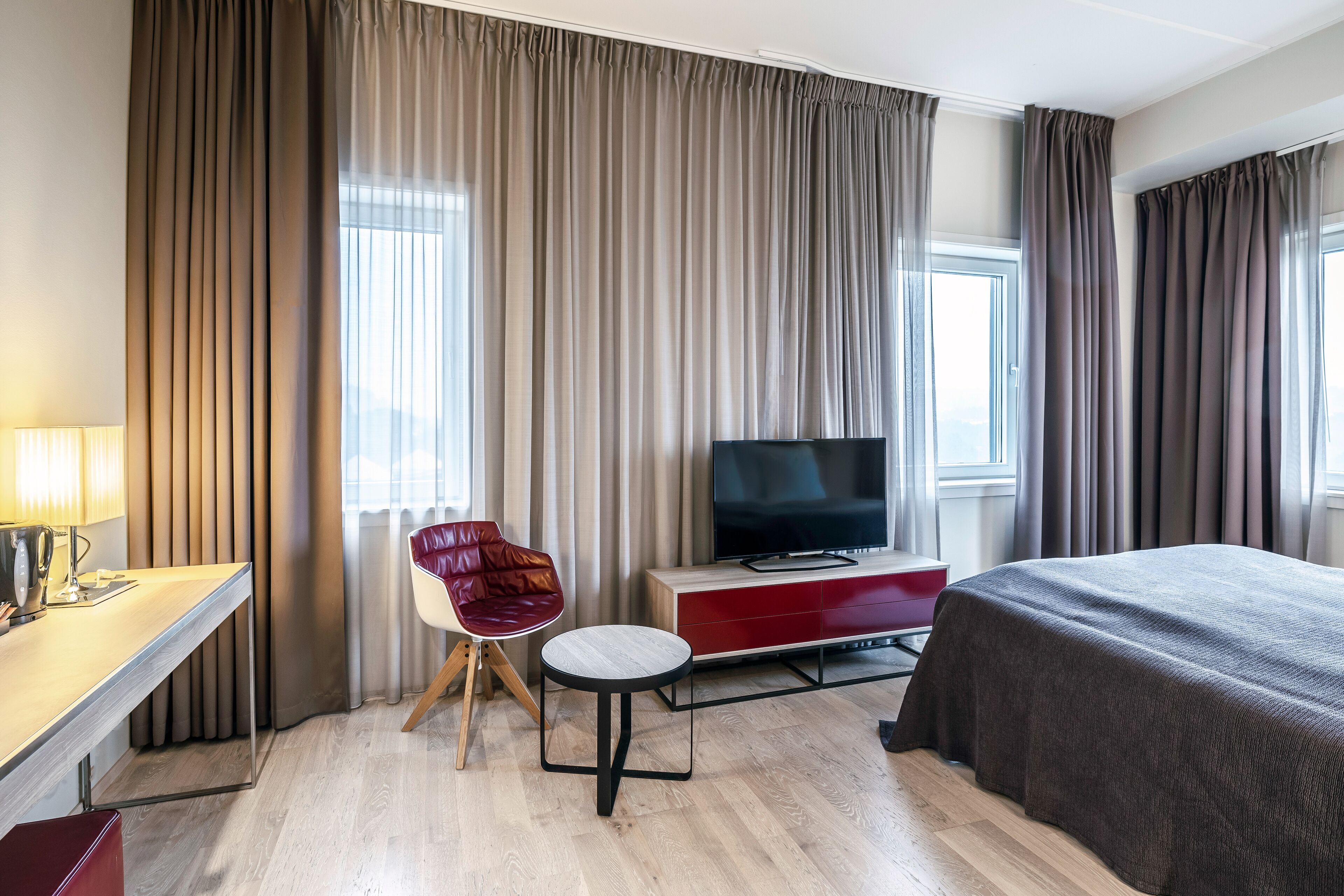 A hotel room with a bed, a TV, a desk with a lamp, a chair, and large windows with curtains.