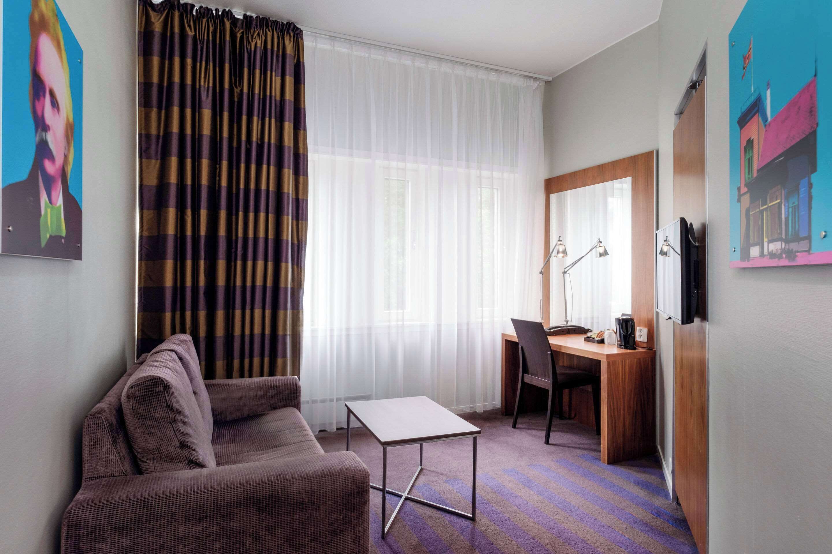 Une chambre d’hôtel avec un canapé, une petite table, un bureau avec une chaise, un miroir et une fenêtre avec des rideaux légers.