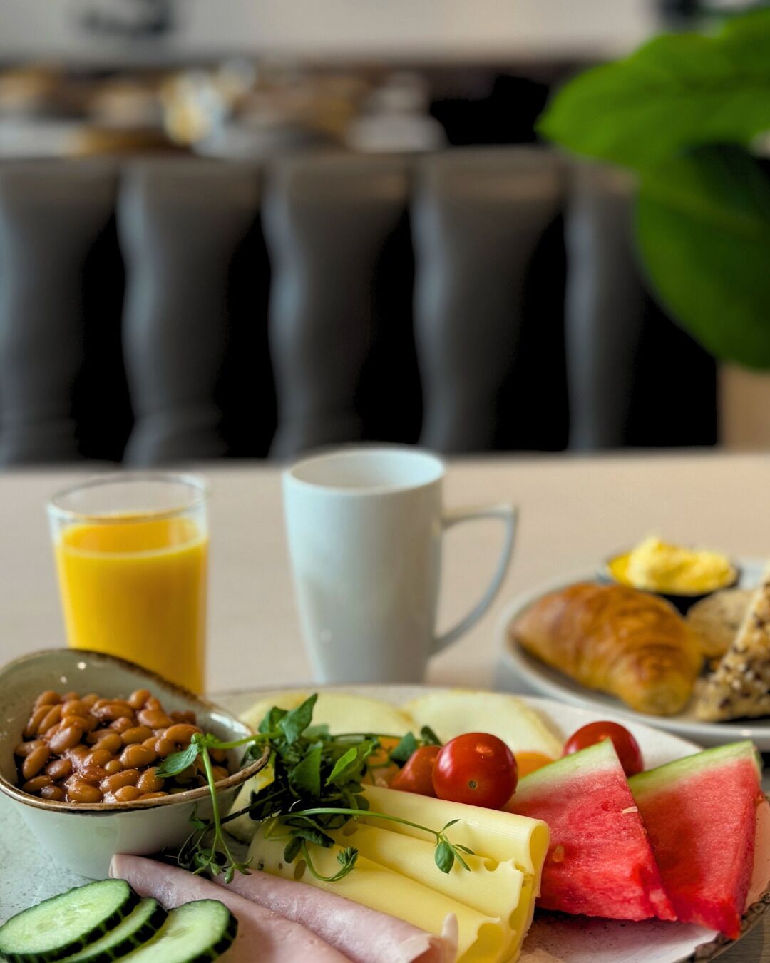 A breakfast setting with a plate of food including beans, cheese, tomatoes, cucumbers, ham, and watermelon, along with a glass of orange juice and a croissant.