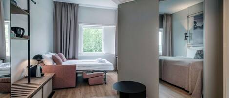 A modern hotel room with a bed, a desk, a chair, and a small table. There is a window with curtains, a door, and a mirror on the wall.
